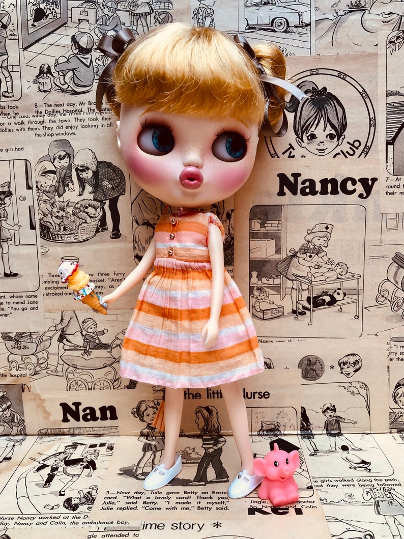 May include: A doll with blonde hair and blue eyes wearing a pink and orange striped dress and white shoes. The doll is holding a yellow and orange bird toy. The doll is standing in front of a vintage magazine page with the text "Nancy" and "the little nurse".