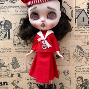 Puede incluir: Una mu&ntilde;eca de estilo vintage vestida con un atuendo de marinero rojo, que incluye una boina con un emblema de ancla. La mu&ntilde;eca tiene cabello oscuro, piel p&aacute;lida y botas negras. El fondo es un collage de tiras c&oacute;micas vintage y conchas marinas.