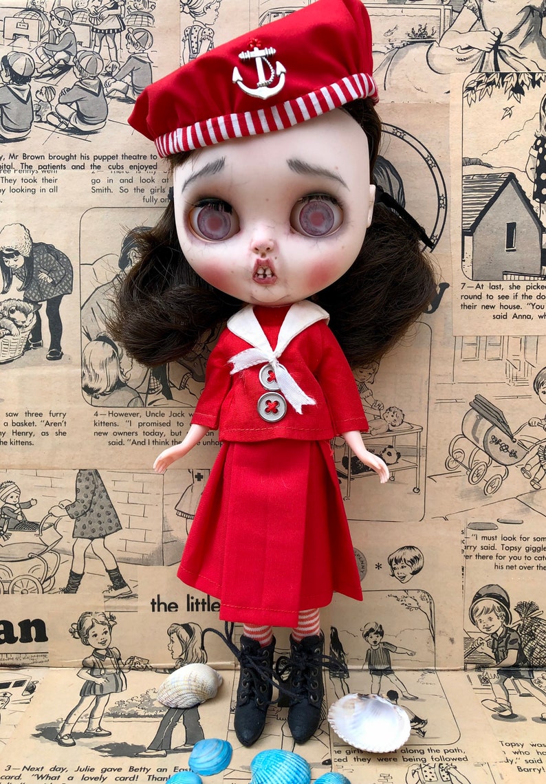 Puede incluir: Una mu&ntilde;eca de estilo vintage vestida con un atuendo de marinero rojo, que incluye una boina con un emblema de ancla. La mu&ntilde;eca tiene cabello oscuro, piel p&aacute;lida y botas negras. El fondo es un collage de tiras c&oacute;micas vintage y conchas marinas.