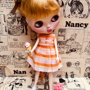 May include: A doll with blonde hair in pigtails wearing a pink and orange striped dress and white shoes. The doll is holding an ice cream cone and a pink elephant toy. The doll is standing in front of a background of vintage comic book pages with the text "Nancy" and "Nan" visible.
