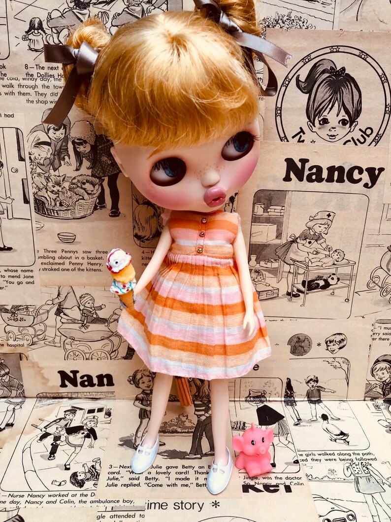 May include: A doll with blonde hair in pigtails wearing a pink and orange striped dress and white shoes. The doll is holding an ice cream cone and a pink elephant toy. The doll is standing in front of a background of vintage comic book pages with the text "Nancy" and "Nan" visible.