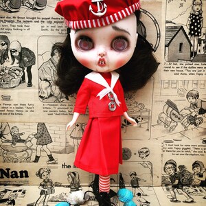Puede incluir: Una mu&ntilde;eca de estilo vintage con cabello oscuro y ojos grandes, vestida con un atuendo de marinero rojo y un sombrero con un emblema de ancla. La mu&ntilde;eca tiene una apariencia &uacute;nica, ligeramente desgastada, sobre un fondo de c&oacute;mics vintage y conchas marinas.