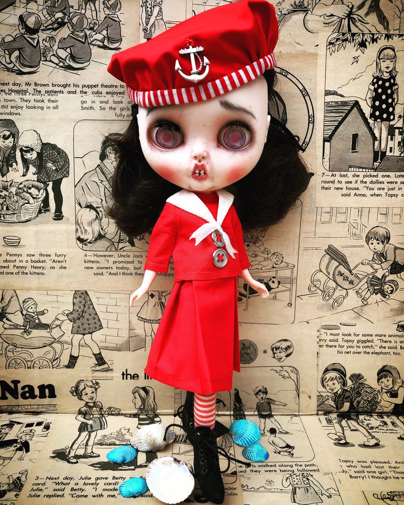 Puede incluir: Una mu&ntilde;eca de estilo vintage con cabello oscuro y ojos grandes, vestida con un atuendo de marinero rojo y un sombrero con un emblema de ancla. La mu&ntilde;eca tiene una apariencia &uacute;nica, ligeramente desgastada, sobre un fondo de c&oacute;mics vintage y conchas marinas.