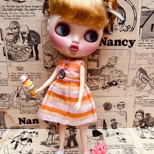May include: A doll with blonde hair wearing a striped orange and white dress and white shoes. The doll is holding an ice cream cone and is standing in front of a pink elephant toy. The background is a collage of vintage paper with text and illustrations.