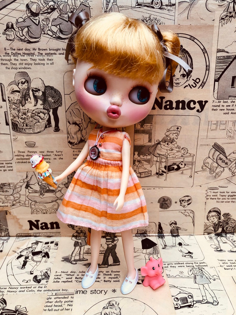 May include: A doll with blonde hair wearing a striped orange and white dress and white shoes. The doll is holding an ice cream cone and is standing in front of a pink elephant toy. The background is a collage of vintage paper with text and illustrations.