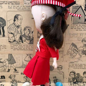 Puede incluir: Una mu&ntilde;eca vestida con un atuendo rojo, que incluye una boina con rayas blancas, una chaqueta y una falda. La mu&ntilde;eca tiene cabello oscuro y lleva botas negras. El fondo presenta tiras c&oacute;micas vintage y conchas marinas.