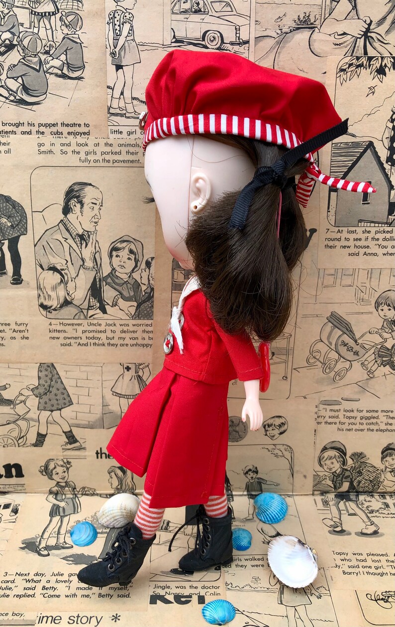 Puede incluir: Una mu&ntilde;eca vestida con un atuendo rojo, que incluye una boina con rayas blancas, una chaqueta y una falda. La mu&ntilde;eca tiene cabello oscuro y lleva botas negras. El fondo presenta tiras c&oacute;micas vintage y conchas marinas.
