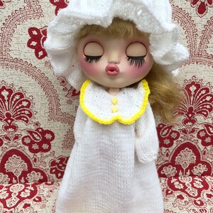 May include: A white knitted doll wearing a white nightgown with yellow trim and a white knitted hat. The doll has blonde hair and closed eyes.