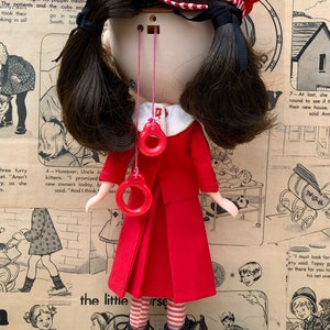 Puede incluir: Mu&ntilde;eca vintage con vestido rojo, sombrero a juego y botas negras. Tiene cabello oscuro y accesorios rojos en forma de anillo. El fondo es un collage de c&oacute;mics vintage y conchas marinas.