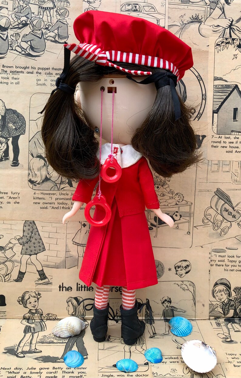 Puede incluir: Mu&ntilde;eca vintage con vestido rojo, sombrero a juego y botas negras. Tiene cabello oscuro y accesorios rojos en forma de anillo. El fondo es un collage de c&oacute;mics vintage y conchas marinas.