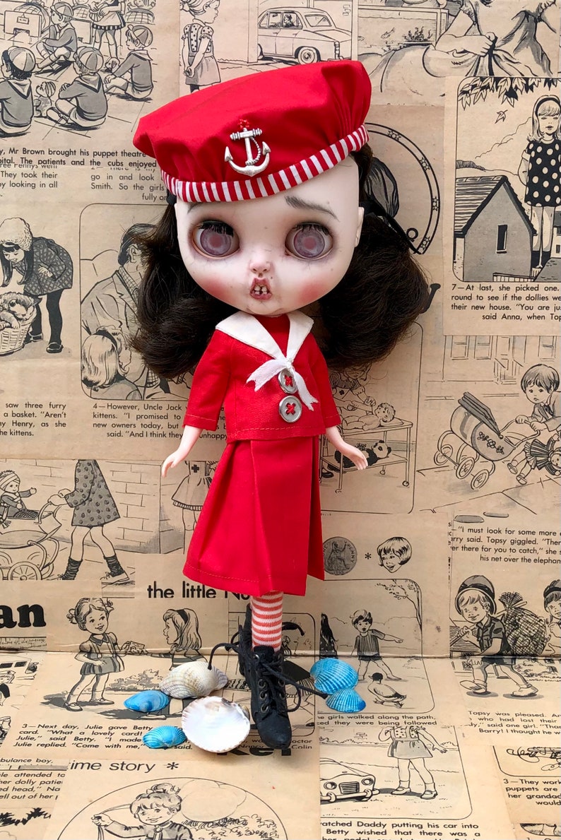 Puede incluir: Mu&ntilde;eca vestida con un atuendo de marinero rojo, que incluye boina con emblema de ancla, chaqueta y falda. La mu&ntilde;eca tiene ojos grandes y posa sobre un fondo de peri&oacute;dico vintage. Botas negras y calcetines a rayas completan el look.