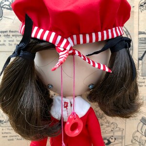Puede incluir: Mu&ntilde;eca vintage con un vestido rojo y boina a juego con una banda a rayas blancas y rojas. La mu&ntilde;eca tiene coletas marrones oscuras y un collar con dos colgantes circulares rojos. Fondo de papel vintage.