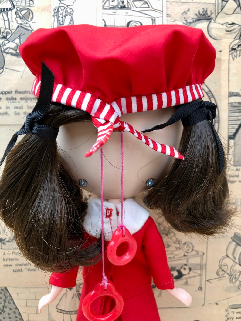 Puede incluir: Mu&ntilde;eca vintage con un vestido rojo y boina a juego con una banda a rayas blancas y rojas. La mu&ntilde;eca tiene coletas marrones oscuras y un collar con dos colgantes circulares rojos. Fondo de papel vintage.