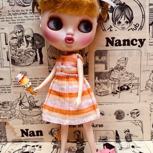 May include: A doll wearing a pink and orange striped dress and white shoes stands in front of a vintage magazine page with the word "Nancy" printed on it. The doll is holding a small orange and white ice cream cone.