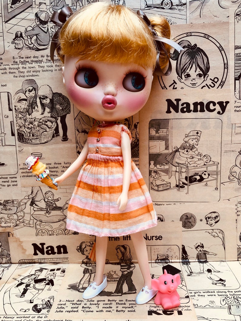 May include: A doll wearing a pink and orange striped dress and white shoes stands in front of a vintage magazine page with the word "Nancy" printed on it. The doll is holding a small orange and white ice cream cone.