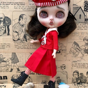 Puede incluir: Una mu&ntilde;eca de estilo vintage con un atuendo de marinero rojo, que incluye una boina con un emblema de ancla. La mu&ntilde;eca tiene cabello oscuro, ojos grandes y posa sobre un fondo de recortes de peri&oacute;dicos antiguos. Botas negras y calcetines a rayas completan el look.