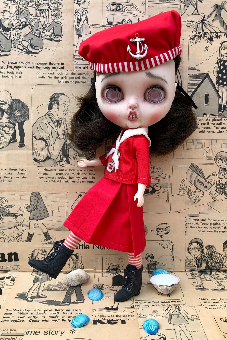 Puede incluir: Una mu&ntilde;eca de estilo vintage con un atuendo de marinero rojo, que incluye una boina con un emblema de ancla. La mu&ntilde;eca tiene cabello oscuro, ojos grandes y posa sobre un fondo de recortes de peri&oacute;dicos antiguos. Botas negras y calcetines a rayas completan el look.