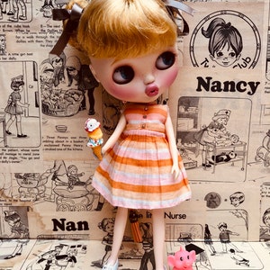 May include: A doll wearing a pink and orange striped dress and white shoes. The doll is holding a small ice cream cone. The doll is standing on a brown surface with a vintage comic book background. The text "Nan" and "e lit Nurse" are visible in the background.