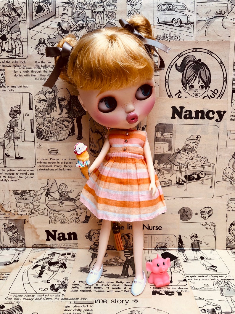 May include: A doll wearing a pink and orange striped dress and white shoes. The doll is holding a small ice cream cone. The doll is standing on a brown surface with a vintage comic book background. The text "Nan" and "e lit Nurse" are visible in the background.