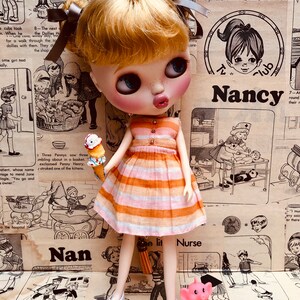 May include: A doll wearing a striped orange and pink dress and white shoes. The doll is holding a pink and white ice cream cone. The doll is standing in front of a background of vintage illustrations and text. The text includes the words "Nancy", "Nan", and "Nurse".