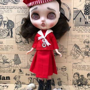 Puede incluir: Mu&ntilde;eca vestida con un atuendo de marinero rojo, que incluye una boina con un emblema de ancla. La mu&ntilde;eca tiene cabello oscuro, piel p&aacute;lida y ojos grandes y expresivos. Hay conchas marinas dispuestas a los pies de la mu&ntilde;eca.