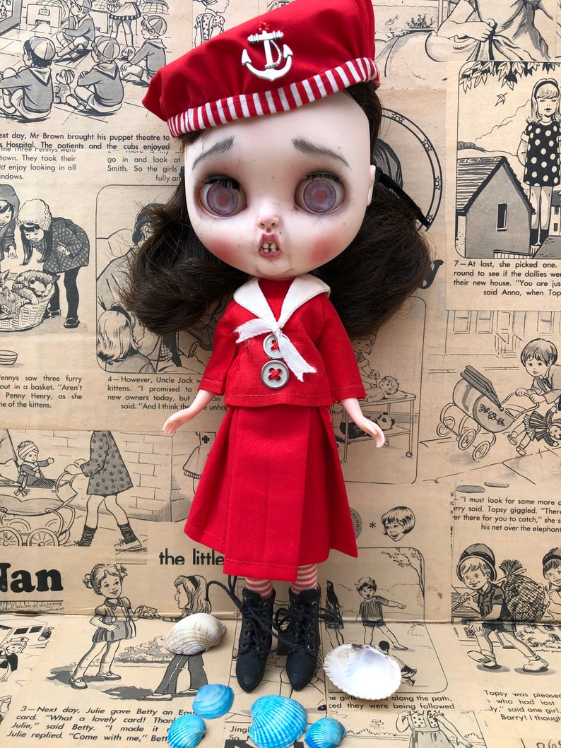 Puede incluir: Mu&ntilde;eca vestida con un atuendo de marinero rojo, que incluye una boina con un emblema de ancla. La mu&ntilde;eca tiene cabello oscuro, piel p&aacute;lida y ojos grandes y expresivos. Hay conchas marinas dispuestas a los pies de la mu&ntilde;eca.