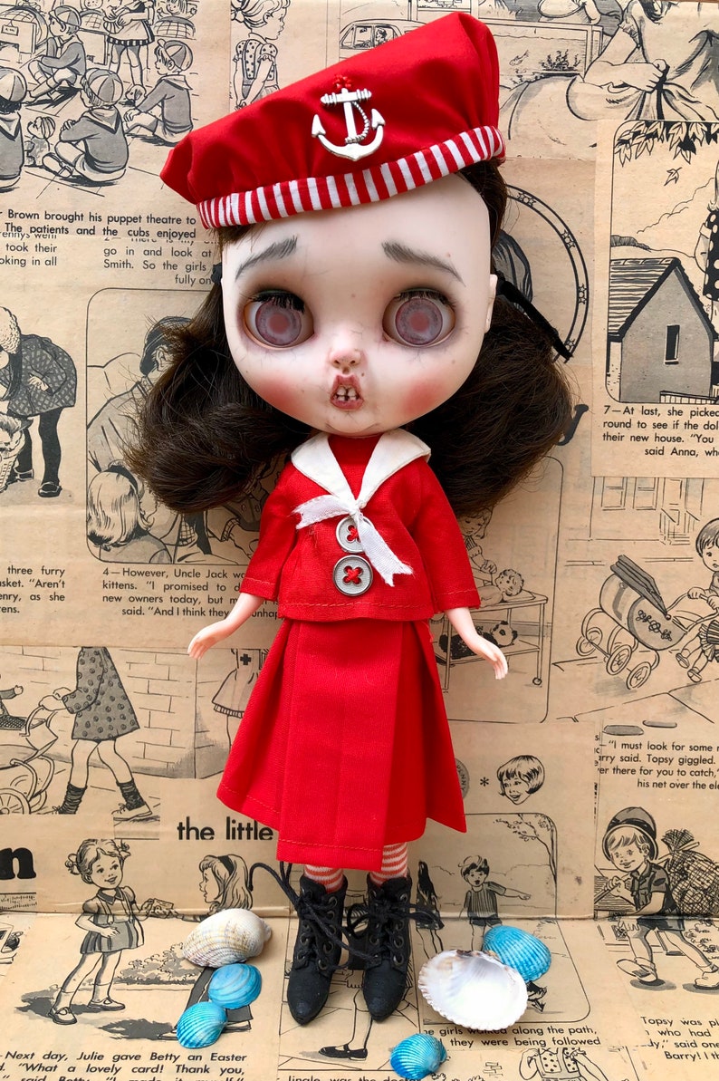 Puede incluir: Mu&ntilde;eca vestida con un atuendo de marinero rojo, que incluye una boina con un emblema de ancla. La mu&ntilde;eca tiene cabello oscuro, piel p&aacute;lida y una expresi&oacute;n ligeramente angustiada. La mu&ntilde;eca lleva botas negras y est&aacute; rodeada de conchas marinas.