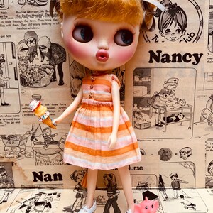 May include: A doll with blonde hair wearing a pink and orange striped dress and white shoes. The doll is holding a pink toy elephant. The doll is standing on a patterned surface with text that includes the word "Nancy".