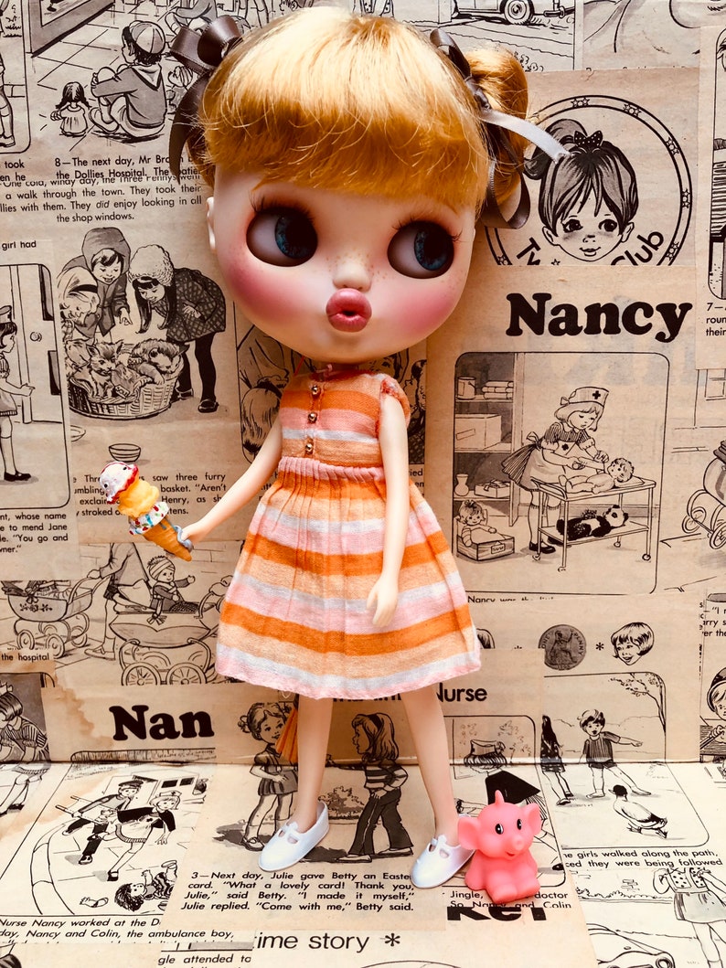 May include: A doll with blonde hair wearing a pink and orange striped dress and white shoes. The doll is holding a pink toy elephant. The doll is standing on a patterned surface with text that includes the word "Nancy".