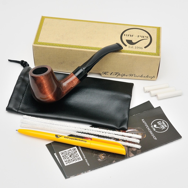 May include: A brown tobacco pipe with a black stem, presented with a black pouch, cleaning tools, and a yellow pen. The pipe is in front of a box with the text "KAF-PIPE Est.1996".