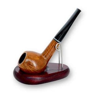 Op de afbeelding: Een houten tabakspijp met een donkerbruine ketel en een zwarte steel, rustend op een kleine, ovale, donkerrode houten standaard. De pijp wordt op zijn plaats gehouden door een zilverkleurige metalen standaard.