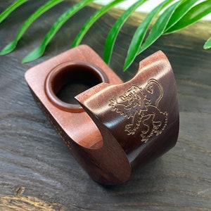May include: A wooden pipe stand with a lion design. The stand is made of dark wood and has a circular opening for the pipe bowl. The lion design is carved into the side of the stand.