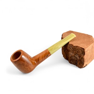 Op de afbeelding: Een gebogen houten tabakspijp met een lichtgekleurd mondstuk. De pijp heeft een warme bruine kleur met een gladde afwerking. De steel heeft de letters "KAF" gegraveerd. Een stuk bruin hout ligt naast de pijp.