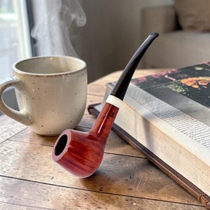 Op de afbeelding: Een gepolijste, roodbruine tabakspijp met een zwarte steel en witte accentring. De pijp ligt op een houten oppervlak naast een kop koffie en een open boek. Stoom stijgt op uit de beker, wat warmte suggereert.