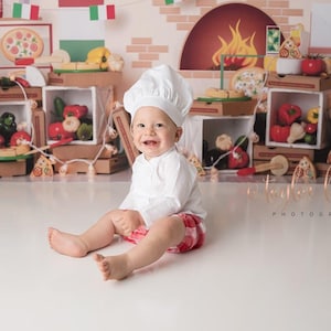 May include: A baby wearing a white chef hat and a white shirt sits on a white surface in front of a backdrop depicting a pizza shop. The backdrop features a brick wall, a pizza oven, and shelves stocked with pizza boxes, vegetables, and pizza slices.