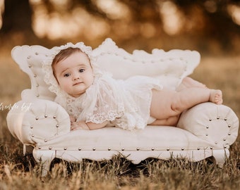 Könnte beinhalten: Ein Baby in einem weißen Spitzenkleid und einer weißen Spitzenhaube liegt auf einem weißen, gepolsterten Sofa. Das Sofa steht auf einem Bett aus braunem Gras.