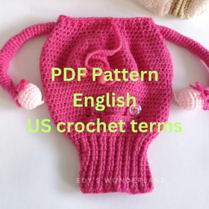Puede incluir: Sombrero de crochet rosa con brazos largos y pequeñas bolas rosas en los extremos. El sombrero tiene un pequeño bolsillo en la parte delantera. El texto "PDF Pattern English US crochet terms" está escrito en el sombrero.