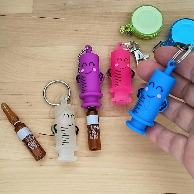 May include: Five colourful syringe-shaped keychains with smiling faces. Each keychain has a small vial attached. The keychains are purple, pink, blue, white, and clear.