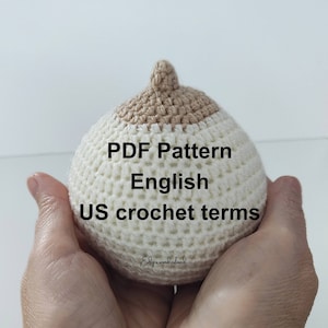 May include: A cream and tan crocheted breast-shaped item. The text "PDF Pattern English US crochet terms" is visible on the item. The item is held in a person's hands.