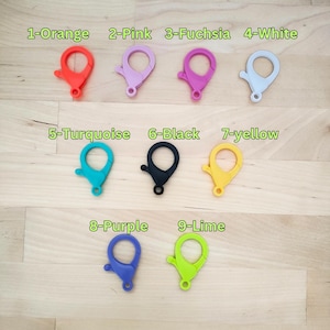 May include: Nine colourful plastic clasps or keychains in various colours, including orange, pink, fuchsia, white, turquoise, black, yellow, purple, and lime green. The clasps are arranged in three rows on a wooden surface.