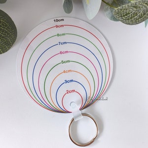 May include: A white circular measuring tool with a keychain ring. The tool has concentric circles with measurements in centimetres from 2 cm to 10 cm. The circles are coloured red, orange, yellow, green, blue, pink, and purple.