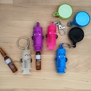 May include: Five colourful syringe-shaped keychains with smiling faces. Each keychain has a small vial attached and a retractable cord. The keychains are on a wooden surface.