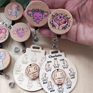 May include: Wooden badge reels with various designs. One reel features a uterus with a floral crown and the word "Cuterus". Another shows a baby held in hands surrounded by flowers. The badge reels have a name tag with the name "Isabella" and "Charlotte".