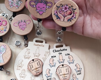 May include: Wooden badge reels with various designs. One reel features a uterus with a floral crown and the word "Cuterus". Another shows a baby held in hands surrounded by flowers. The badge reels have a name tag with the name "Isabella" and "Charlotte".