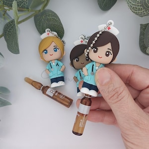 May include: Three nurse-themed keychains with different skin tones and hair colors. Each keychain features a miniature nurse figurine wearing a light blue uniform and a white cap. Two brown glass vials are also visible.