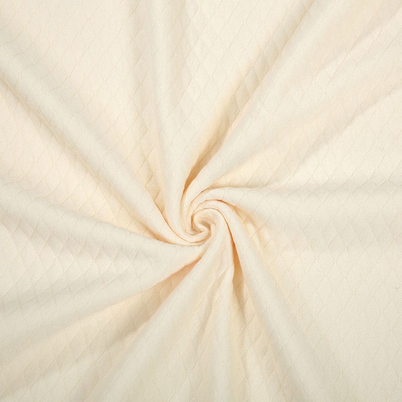 May include: Cream-colored fabric with a diamond pattern, arranged in a spiral. The fabric is soft and appears to be a knit or woven material. The texture is visible, with the diamond pattern adding visual interest to the textile.