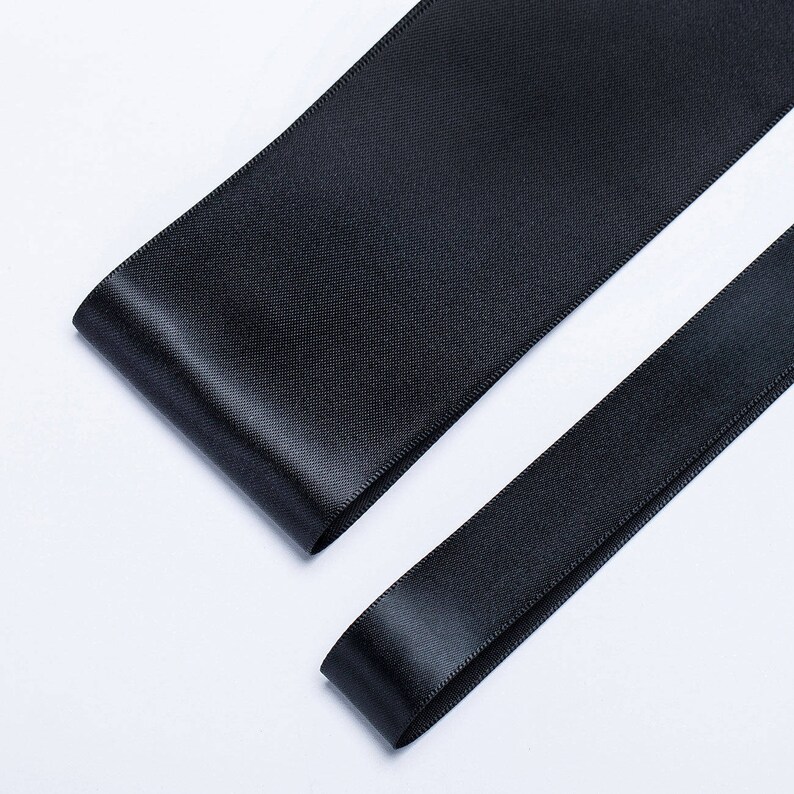 May include: Two black satin ribbons, one wider than the other, are laid out on a white surface.