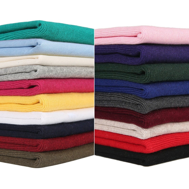 Op de afbeelding: Een stapel van 20 verschillende gekleurde gebreide stofstalen. De stoffen zijn allemaal geribbeld en hebben een zachte textuur. De kleuren omvatten roze, groen, blauw, wit, grijs, rood, geel, zwart en bruin.