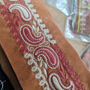 Może przedstawiać: Zbliżenie brązowego paska skórzanego z misternymi białymi i czerwonymi haftami. Wzór zawiera wzory paisley i motywy kwiatowe.