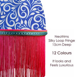 Può includere: Primo piano di un paralume in tessuto con un motivo floreale blu e bianco. Il paralume ha un bordo rosa e una frangia rossa profonda 15 cm. Il testo recita "Neotrims Silky Loop Fringe" e "12 Colori."